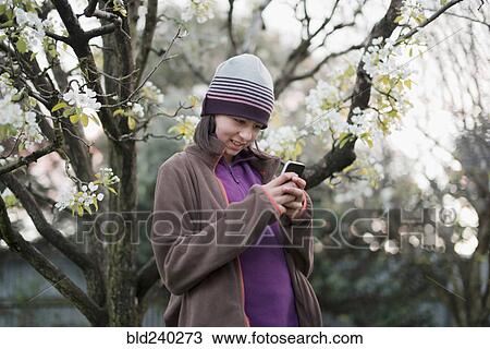Stock Image - Smiling Mixed Race girl texting on cell phone near tree. Fotosearch