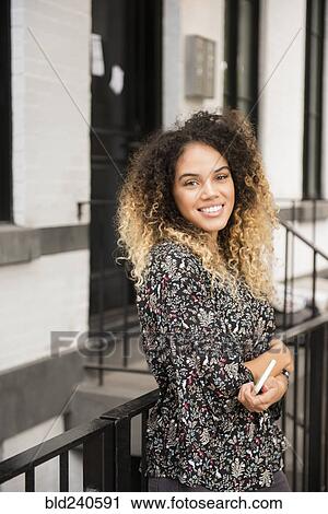 Stock Image - Smiling Mixed Race woman holding cell phone in city. Fotosearch