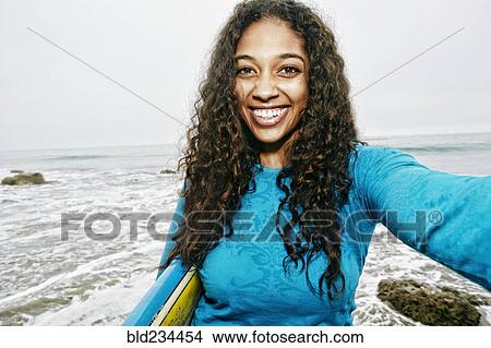 Picture - Smiling Mixed Race woman holding surfboard posing for selfie at beach . Fotosearch