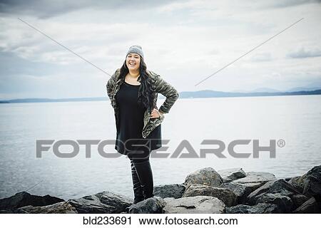 Stock Image - Smiling Mixed Race woman posing on rocks near ocean. Fotosearch
