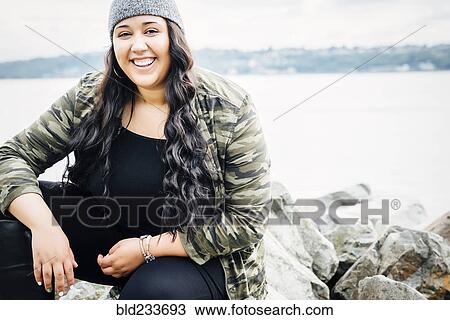 Stock Image - Smiling Mixed Race woman sitting on rocks near ocean. Fotosearch