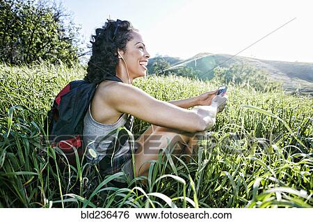 Stock Photograph - Smiling Mixed Race woman sitting on hill listening to cell phone. Fotosearch