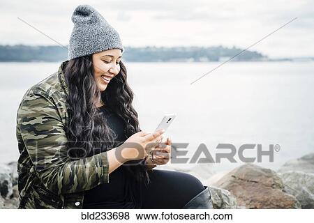 Stock Photo - Smiling Mixed Race woman texting on cell phone near ocean. Fotosearch