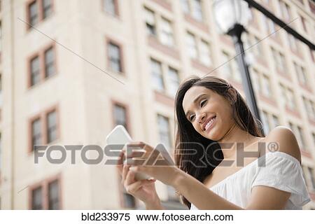 Stock Photography - Smiling Mixed Race woman texting on cell phone in city. Fotosearch