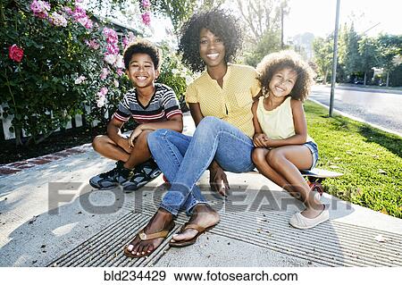 Stock Photo - Smiling mother and children sitting on skateboard on sidewalk. Fotosearch