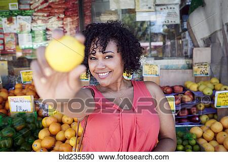 Stock Image - Smiling woman showing lemon at fruit stand. Fotosearch