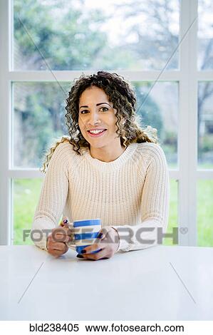 Stock Photography - Smiling woman sitting at table holding coffee cup. Fotosearch