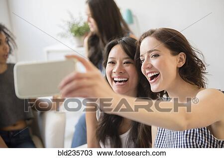 Stock Image - Smiling women posing for cell phone selfie at party. Fotosearch