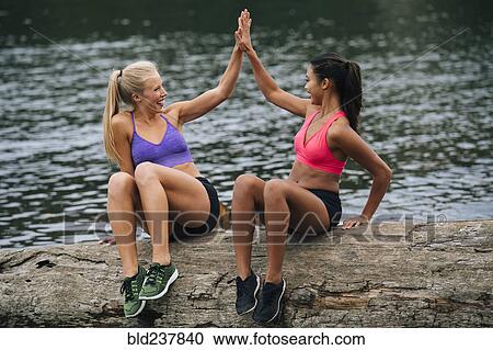 Smiling women sitting on log near lake high-fiving View Large Photo Image Stock Image - Smiling women sitting on log near lake high-fiving. Fotosearch