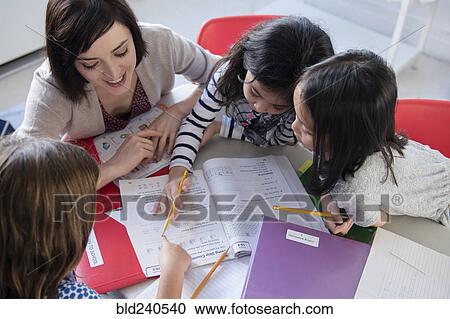 Stock Image - Teacher helping girls with workbook in classroom. Fotosearch