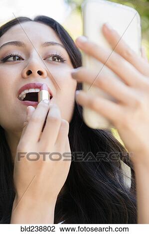 Stock Image - Thai woman holding cell phone applying lipstick. Fotosearch