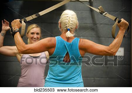 Stock Image - Trainer assisting older woman working out in gymnasium. Fotosearch