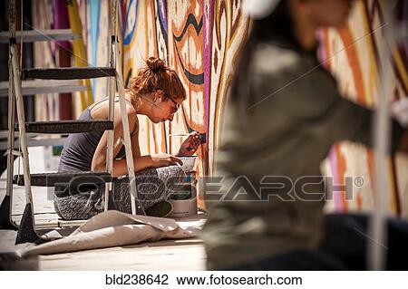 Stock Image - Women sitting on tarp painting mural on wall. Fotosearch