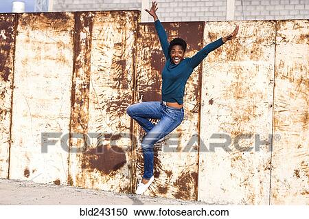 Stock Image - African American woman jumping for joy near rusty metal wall. Fotosearch