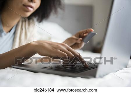 Picture - African American woman laying in bed online shopping with laptop. Fotosearch