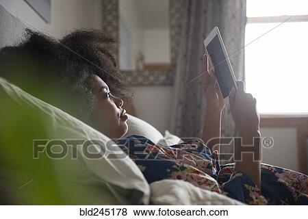 Stock Photo - African American woman laying in bed reading digital tablet. Fotosearch
