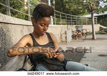 Stock Photo - African American woman on bench at park texting on cell phone. Fotosearch
