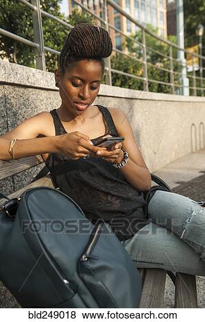 Stock Photo - African American woman on bench at park texting on cell phone. Fotosearch