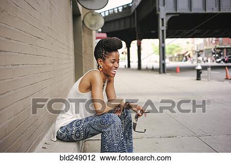 Stock Image - African American woman with braids sitting on ledge and laughing. Fotosearch