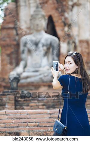 Stock Photo - Asian woman photographing Buddha statue with cell phone. Fotosearch