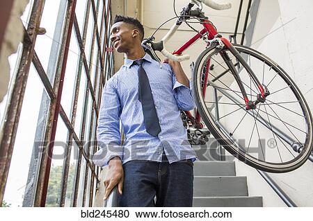 Stock Image - Black businessman descending staircase carrying bicycle. Fotosearch