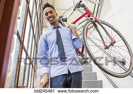 Stock Image - Black businessman descending staircase carrying bicycle. Fotosearch