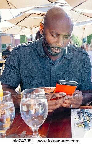 Stock Image - Black man sitting at restaurant table texting on cell phone. Fotosearch