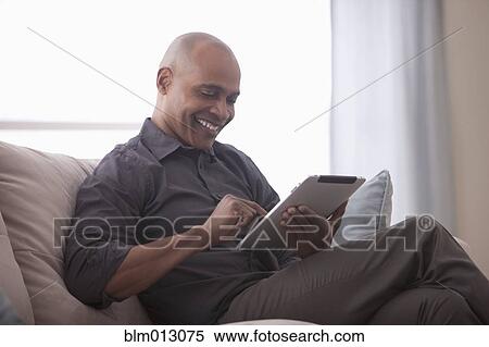 Black man sitting on sofa using digital tablet View Large Photo Image Stock Photography - Black man sitting on sofa using digital tablet. Fotosearch