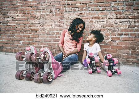 Stock Image - Black mother and daughter wearing roller skates sitting on sidewalk. Fotosearch