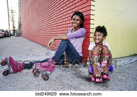 Stock Photo - Black mother and daughter wearing roller skates sitting on sidewalk. Fotosearch