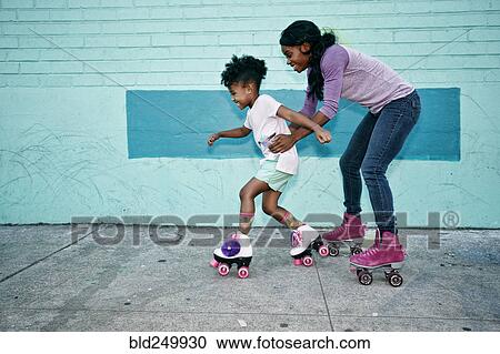 Stock Image - Black mother holding waist of daughter wearing roller skates. Fotosearch