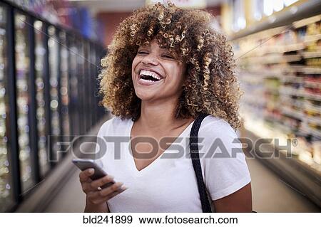 Stock Photo - Black woman holding cell phone laughing in grocery store. Fotosearch