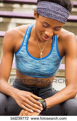 Stock Image - Black woman sitting on bleachers checking smart watch. Fotosearch