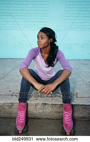 Stock Image - Black woman wearing roller skates sitting on curb. Fotosearch