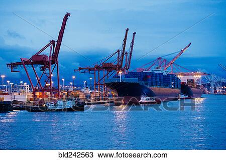 Stock Image - Cargo containers on freighter at shipping port near tugboats. Fotosearch