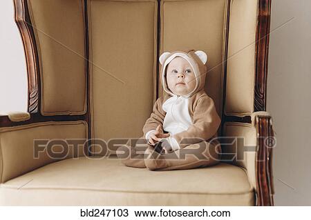 Stock Image - Caucasian baby boy sitting in armchair wearing bear costume. Fotosearch