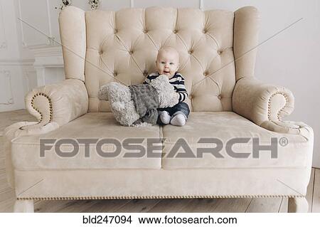 Picture - Caucasian baby boy sitting on love seat playing with teddy bear. Fotosearch