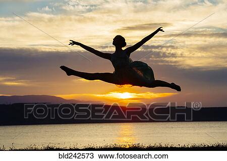 Stock Image - Caucasian ballerina jumping on beach at sunset. Fotosearch