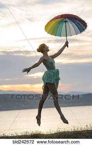Stock Image - Caucasian ballerina jumping with multicolor umbrella on beach. Fotosearch