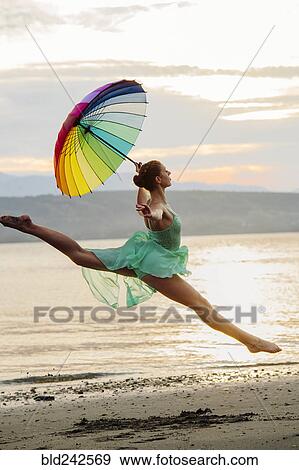 Stock Photo - Caucasian ballerina jumping with multicolor umbrella on beach. Fotosearch