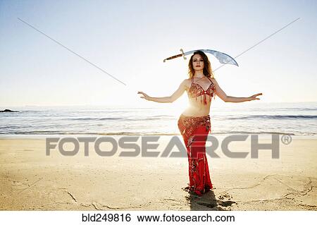 Stock Photograph - Caucasian belly dancer on beach balancing sword on head. Fotosearch