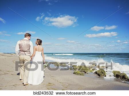 Stock Image - Caucasian bride and groom walking on beach. Fotosearch