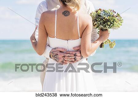 Stock Photo - Caucasian bride and groom with tattoos hugging on beach. Fotosearch