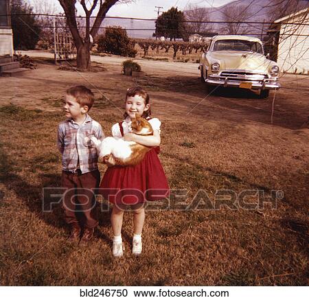 Stock Image - Caucasian brother and sister standing in yard holding cat. Fotosearch