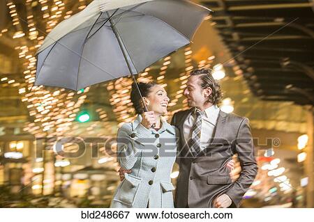 Stock Photo - Caucasian couple carrying umbrella in city at night. Fotosearch