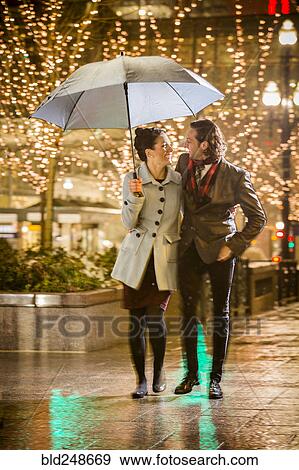 Stock Photo - Caucasian couple carrying umbrella in city at night. Fotosearch