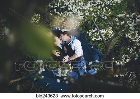 Stock Image - Caucasian couple laying in grass under flowering trees. Fotosearch