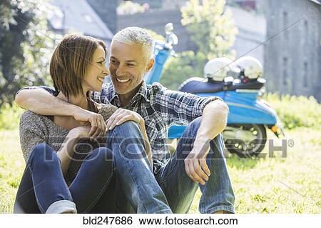 Stock Photograph - Caucasian couple sitting in grass near motor scooter. Fotosearch