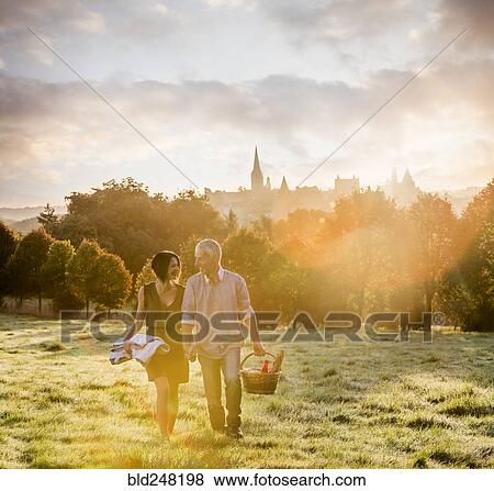 Stock Photo - Caucasian couple walking in field carrying picnicblanket and basket. Fotosearch