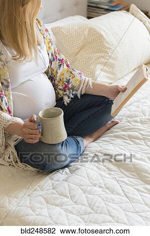 Stock Image - Caucasian expectant mother sitting on bed reading book and drinking coffee. Fotosearch
